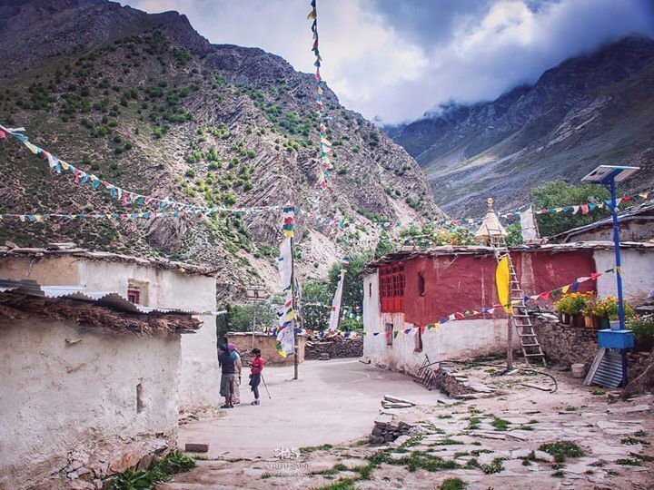 A mountainous village with traditional houses, prayer flags, and a small shrine.