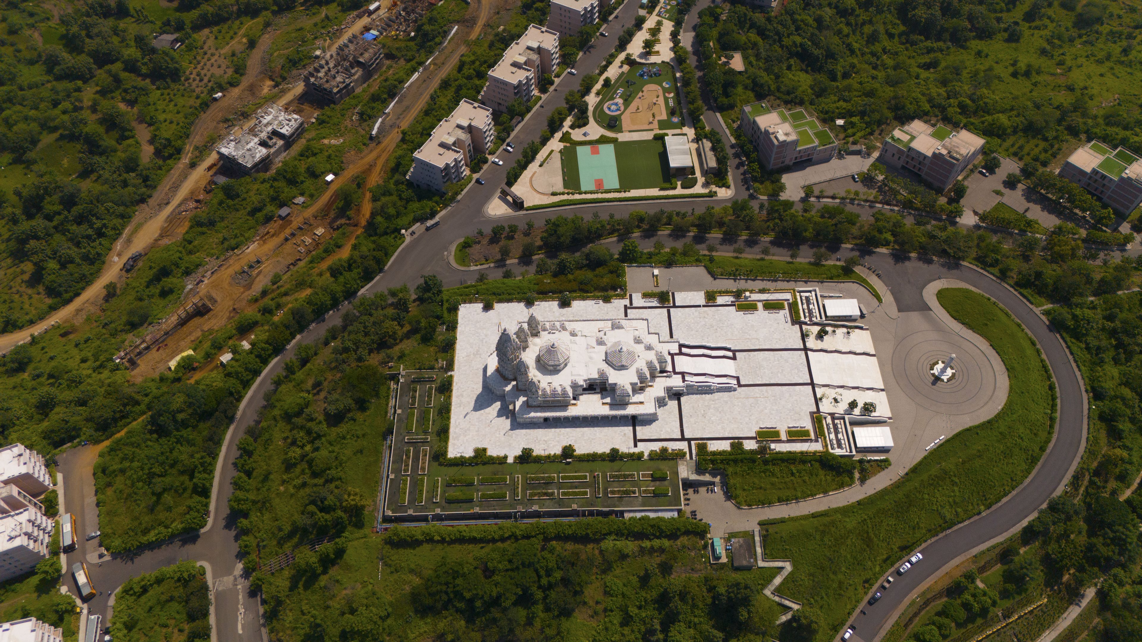 Aerial view of the white marble BAPS Shri Swaminarayan Mandir temple complex surrounded by lush green landscapes, paved walkways, and modern residential buildings in Chikhli, Gujarat, India.