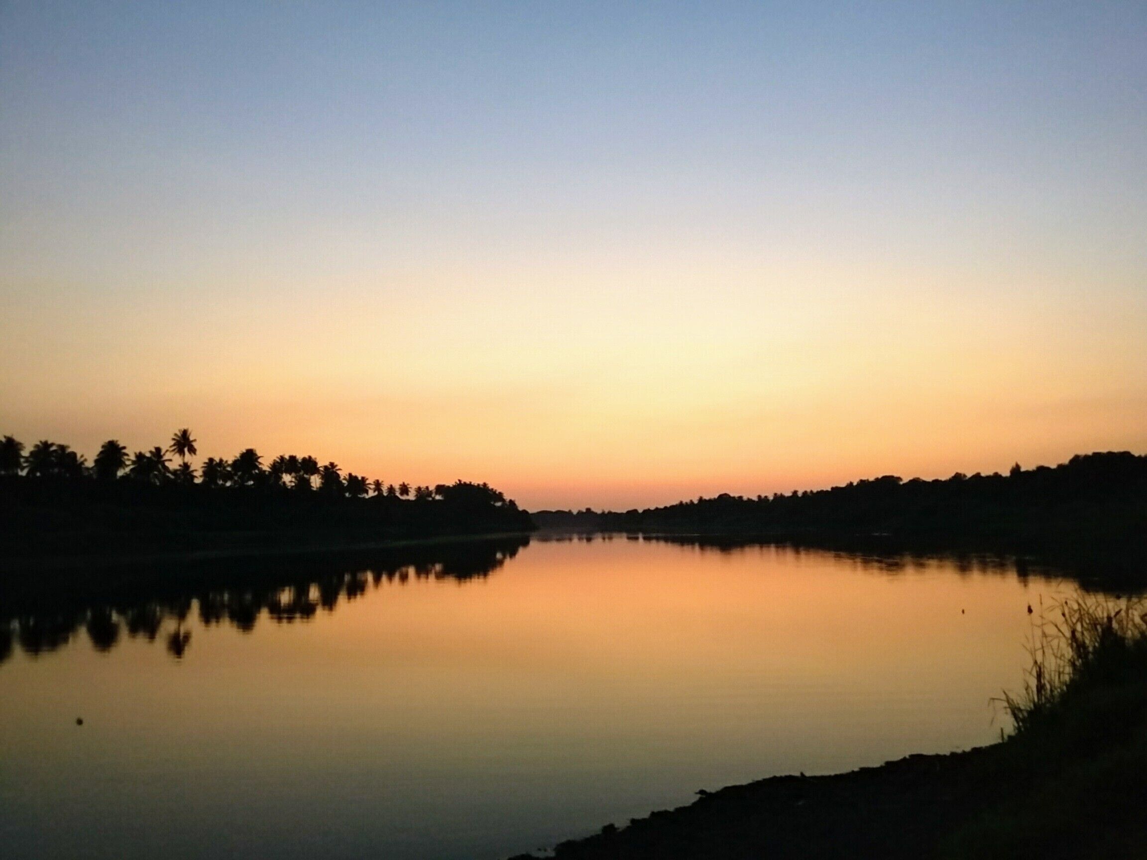 A calm lake at sunset with silhouettes of palm trees and a clear sky.