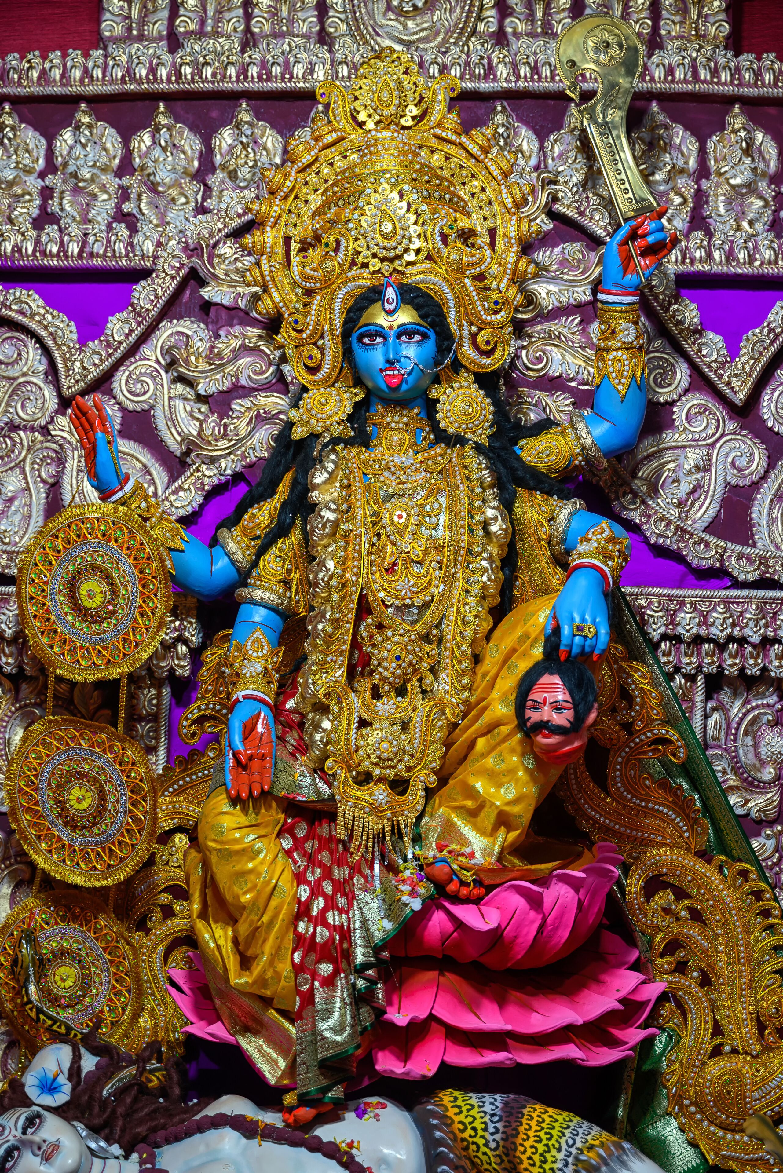 A statue of a deity with intricate golden jewelry and a large headdress, set against a richly decorated background.
