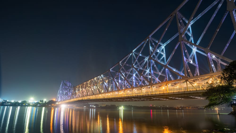 A nighttime view of a large, illuminated bridge with a complex truss structure spanning over a body of water.