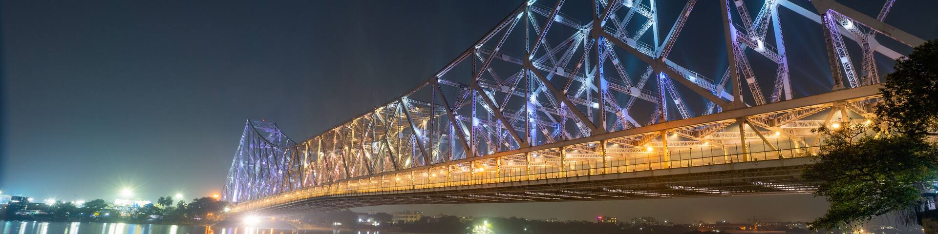 A nighttime view of a large, illuminated bridge with a complex truss structure spanning over a body of water.