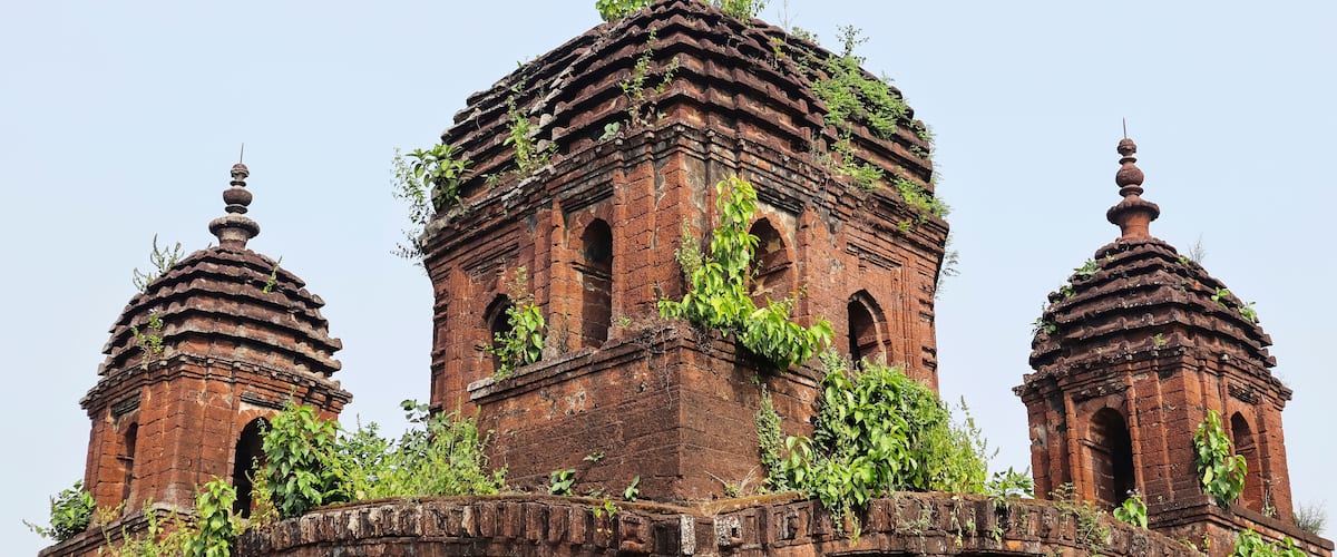 A historical brick structure with three domes, overgrown with vegetation.