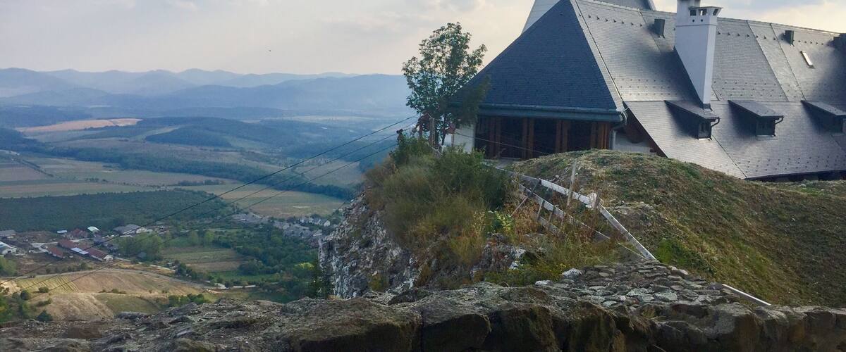 A church with a steep roof and crosses on the roof, overlooking a valley with fields and distant mountains.