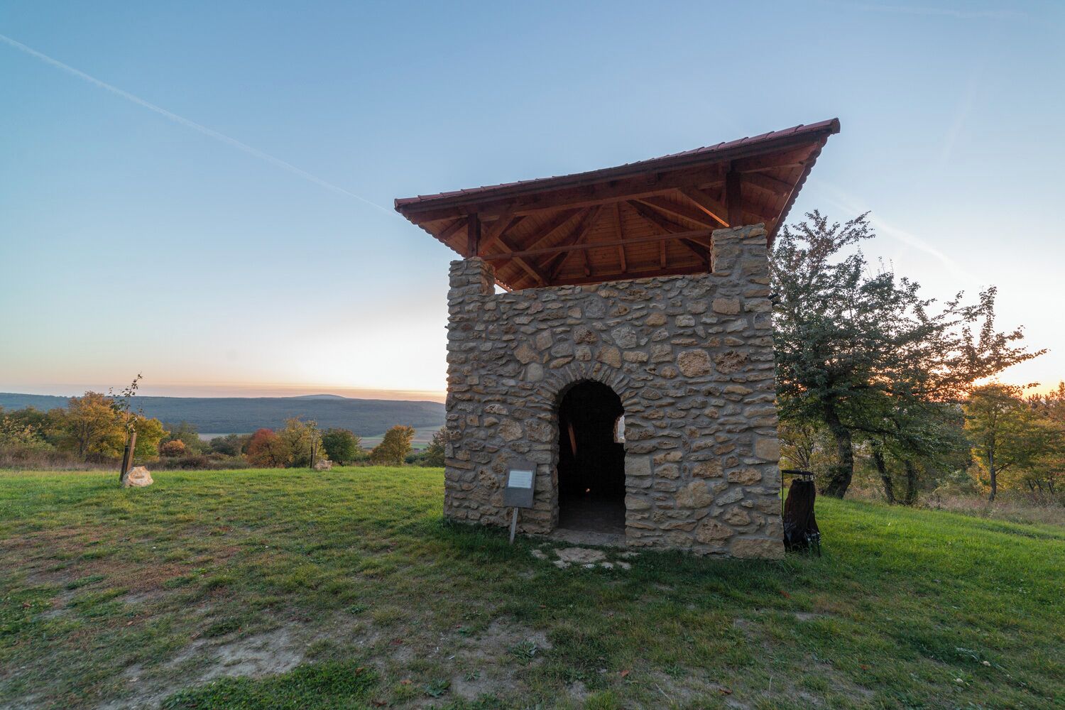 A stone structure with a wooden roof and an arched entrance, surrounded by grass and trees, with a view of distant hills.