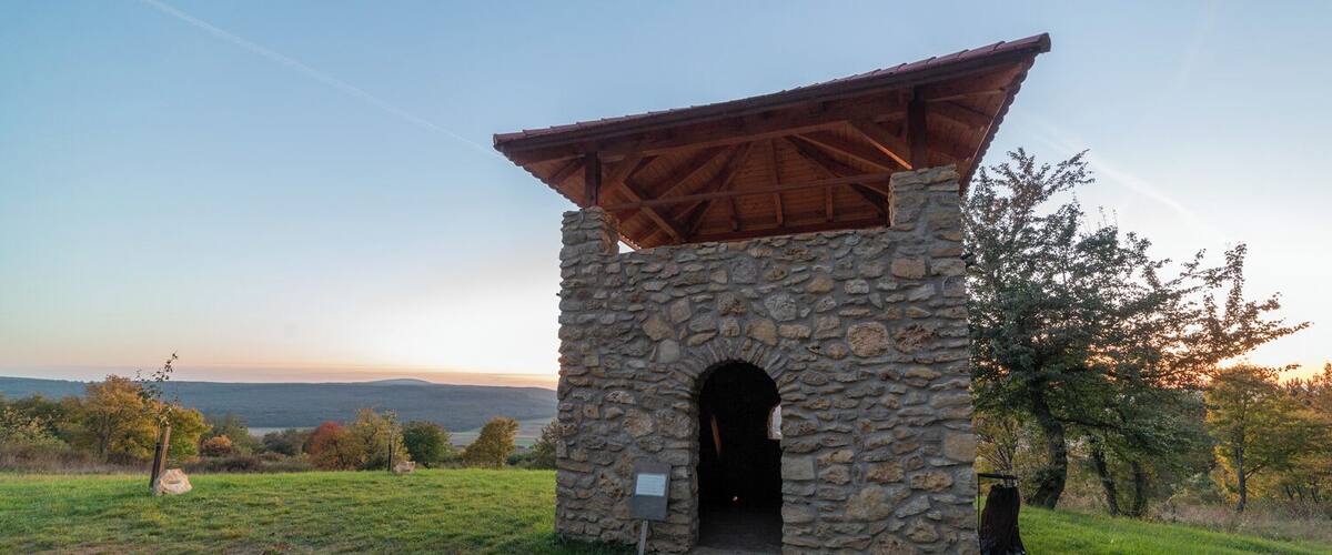 A stone structure with a wooden roof and an arched entrance, surrounded by grass and trees, with a view of distant hills.