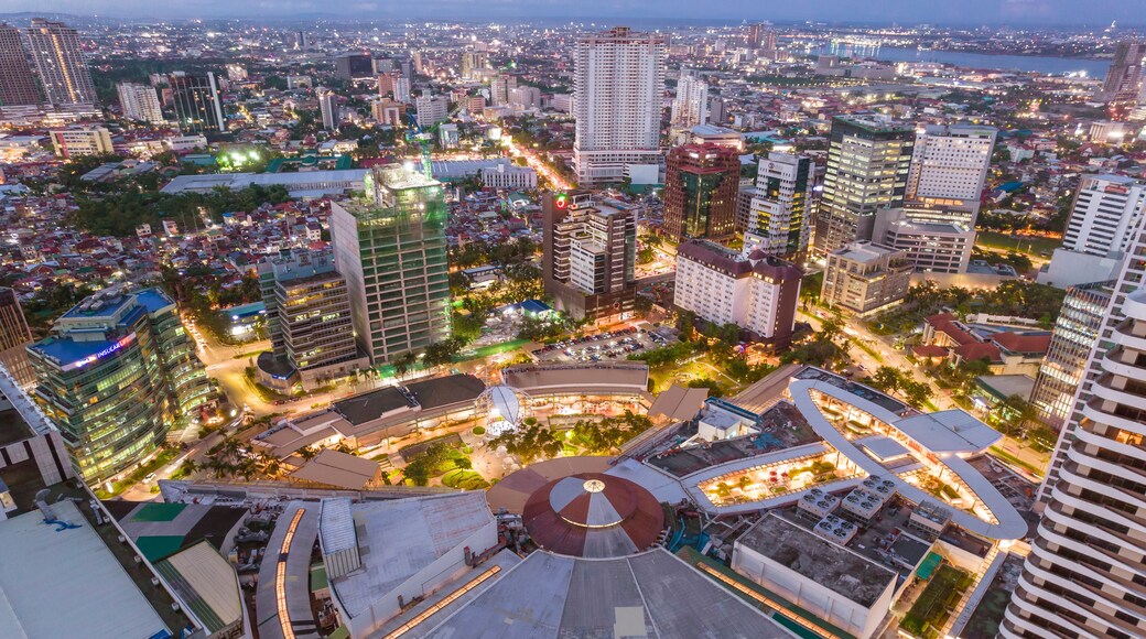 A cityscape at dusk with illuminated buildings, a large circular structure, and a body of water in the distance.