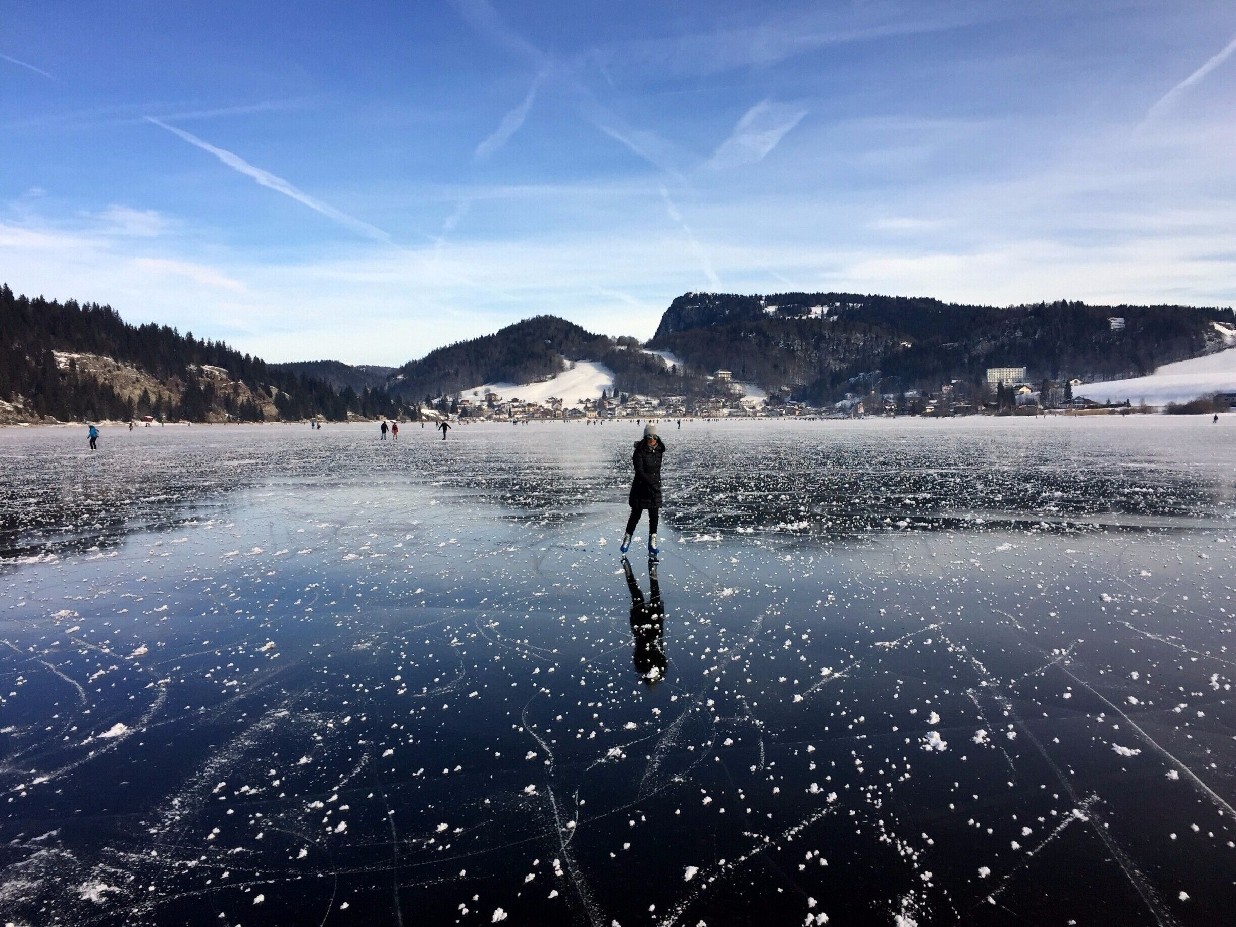 A frozen lake with people ice skating, surrounded by hills and a town in the background.