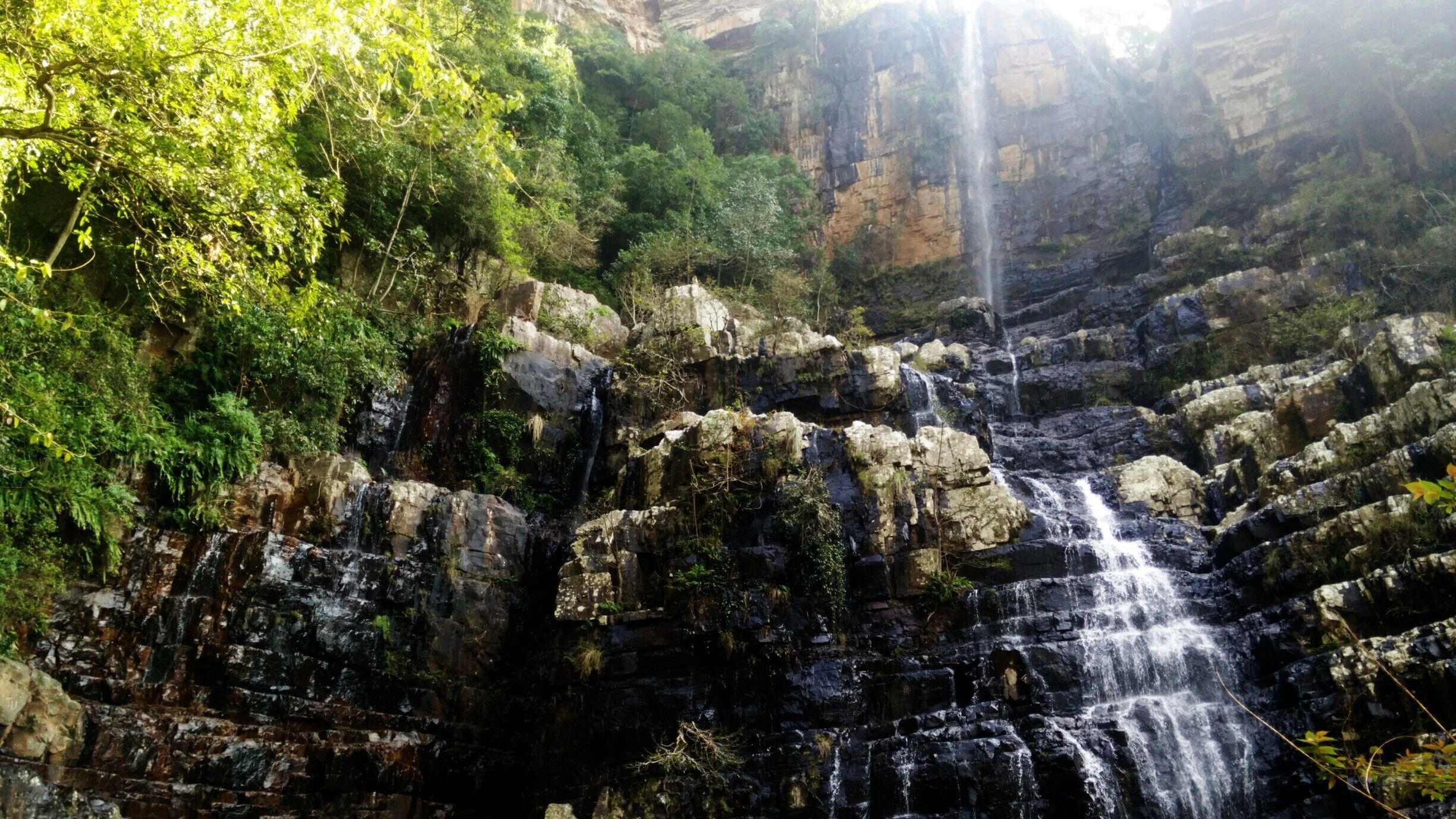 A multi-tiered waterfall surrounded by rocky terrain and dense greenery.