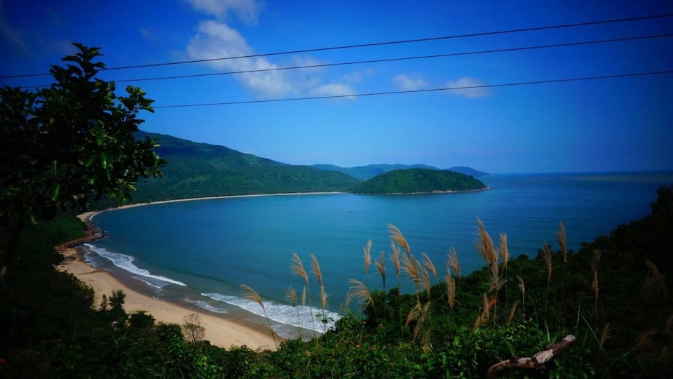A coastal landscape with a sandy beach, calm sea, and lush green hills.