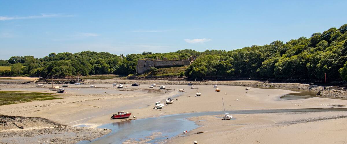 A coastal landscape with a sandy beach, a small river, and a historic stone structure in the background.
