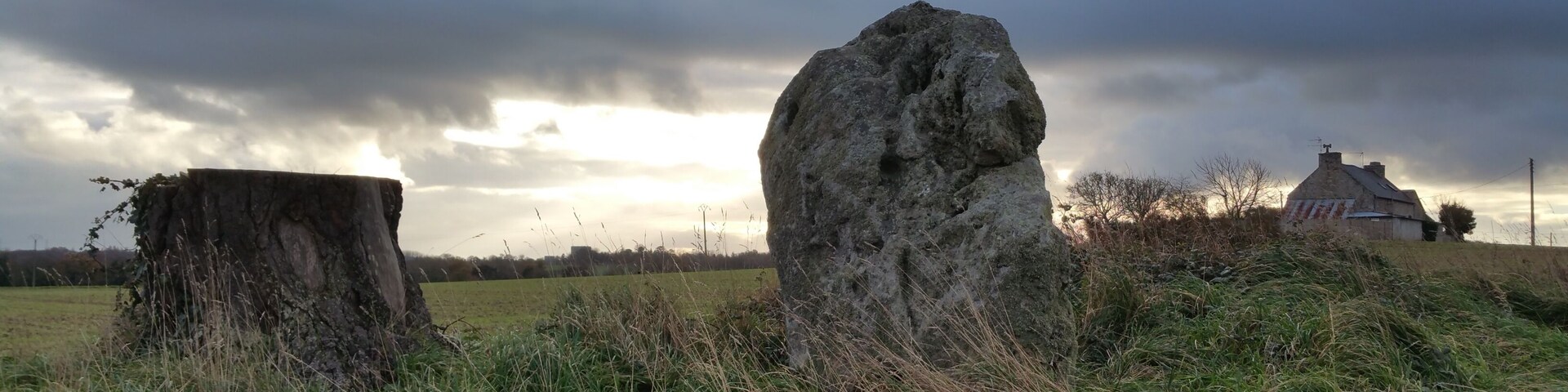A rural landscape with a large standing stone, a tree stump, and a house in the background.