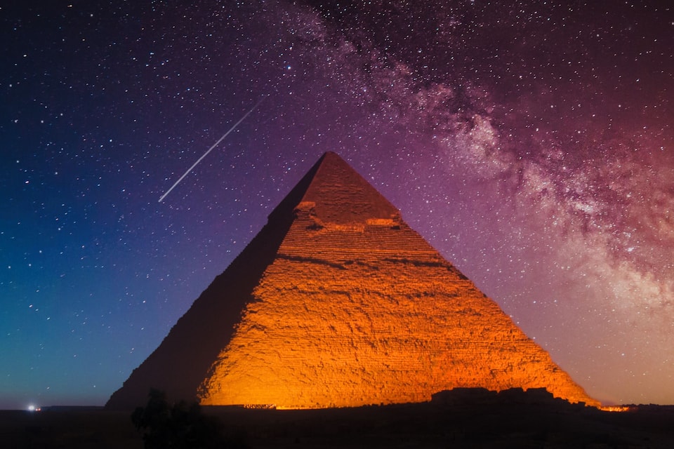Een piramide verlicht in de nacht, met een sterrenhemel en de Melkweg zichtbaar.