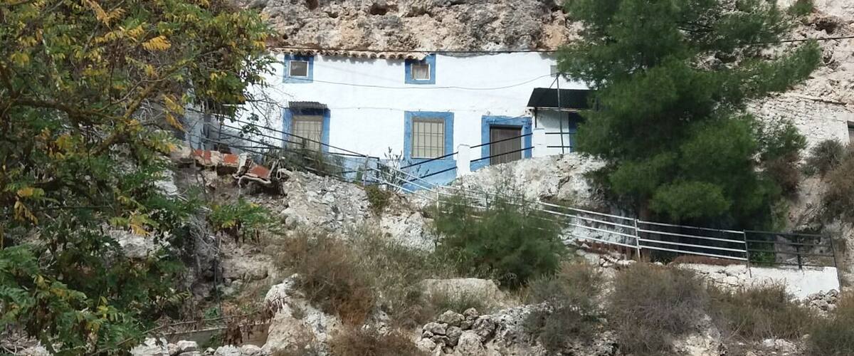 A white building with blue trim nestled against a rocky cliff.