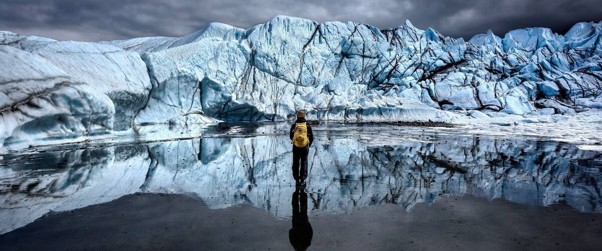 A person standing in front of a glacier with a reflection in the water.