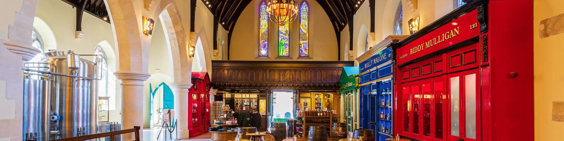 A spacious interior with high ceilings, arched windows, and a central stained glass window. There are wooden tables and stools, and a large barrel labeled 'Pearse Irish Whiskey'.