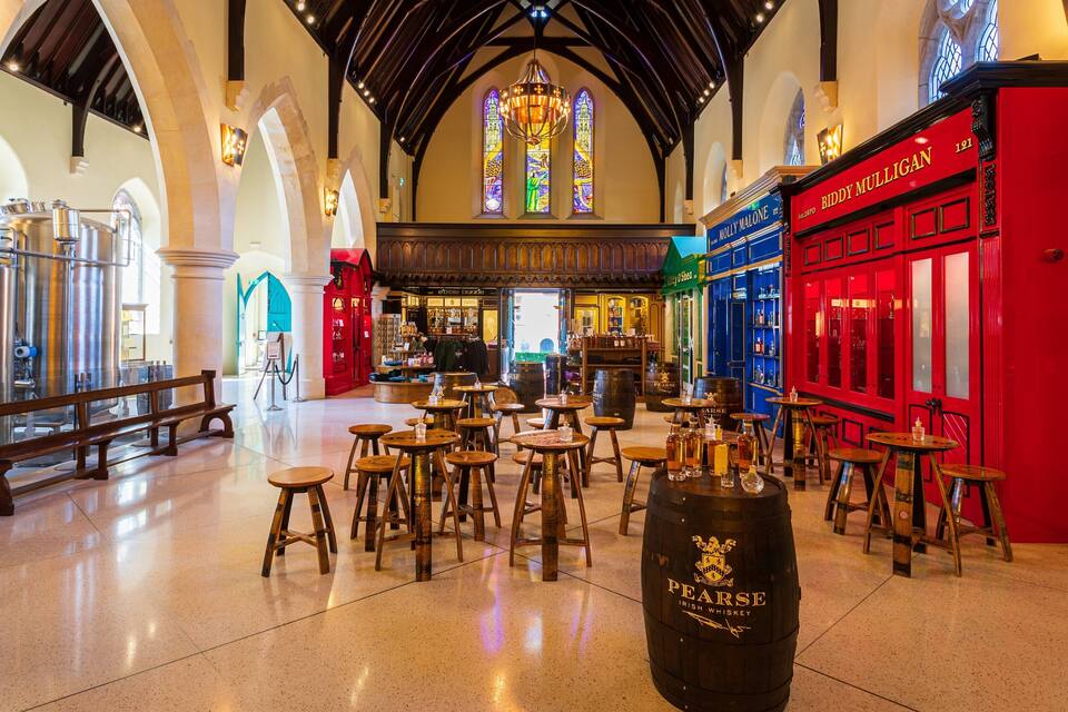 Un interior espacioso con techos altos, ventanas arqueadas y un ventanal central de vidrio emplomado. Hay mesas y taburetes de madera, y un barril grande con la etiqueta 'Pearse Irish Whiskey'.