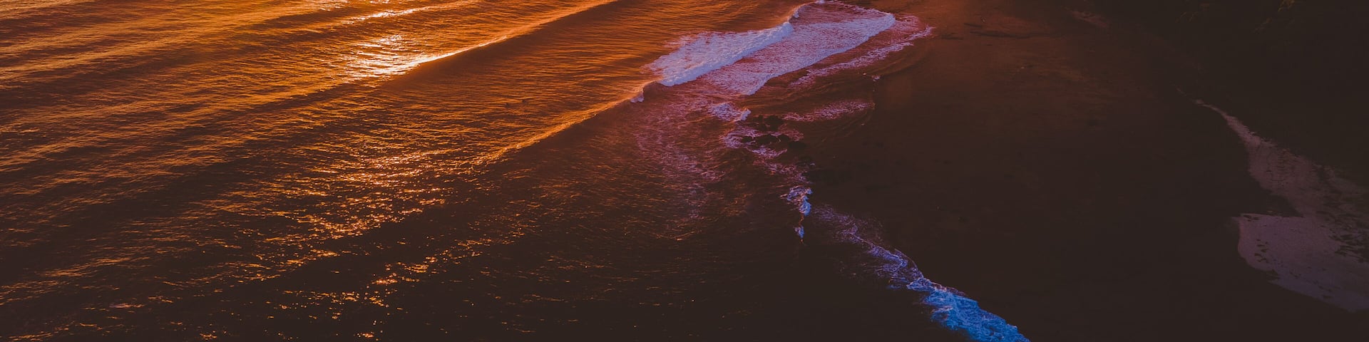 A sunset over a coastal landscape with waves crashing onto the shore and a cliff in the background.