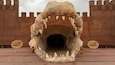 Une grande sculpture réaliste représentant la gueule d’un dinosaure, avec ses dents, placée sur un fond évoquant un château.