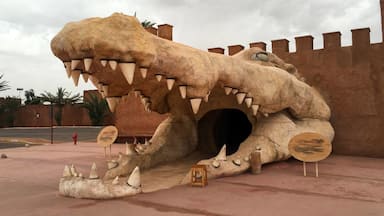 A large dinosaur sculpture with a cave entrance, surrounded by palm trees and a castle-like wall.