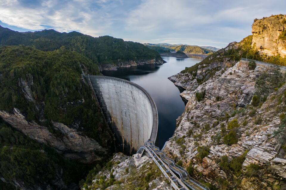 A large dam with a water reservoir, surrounded by forested hills and rocky terrain.