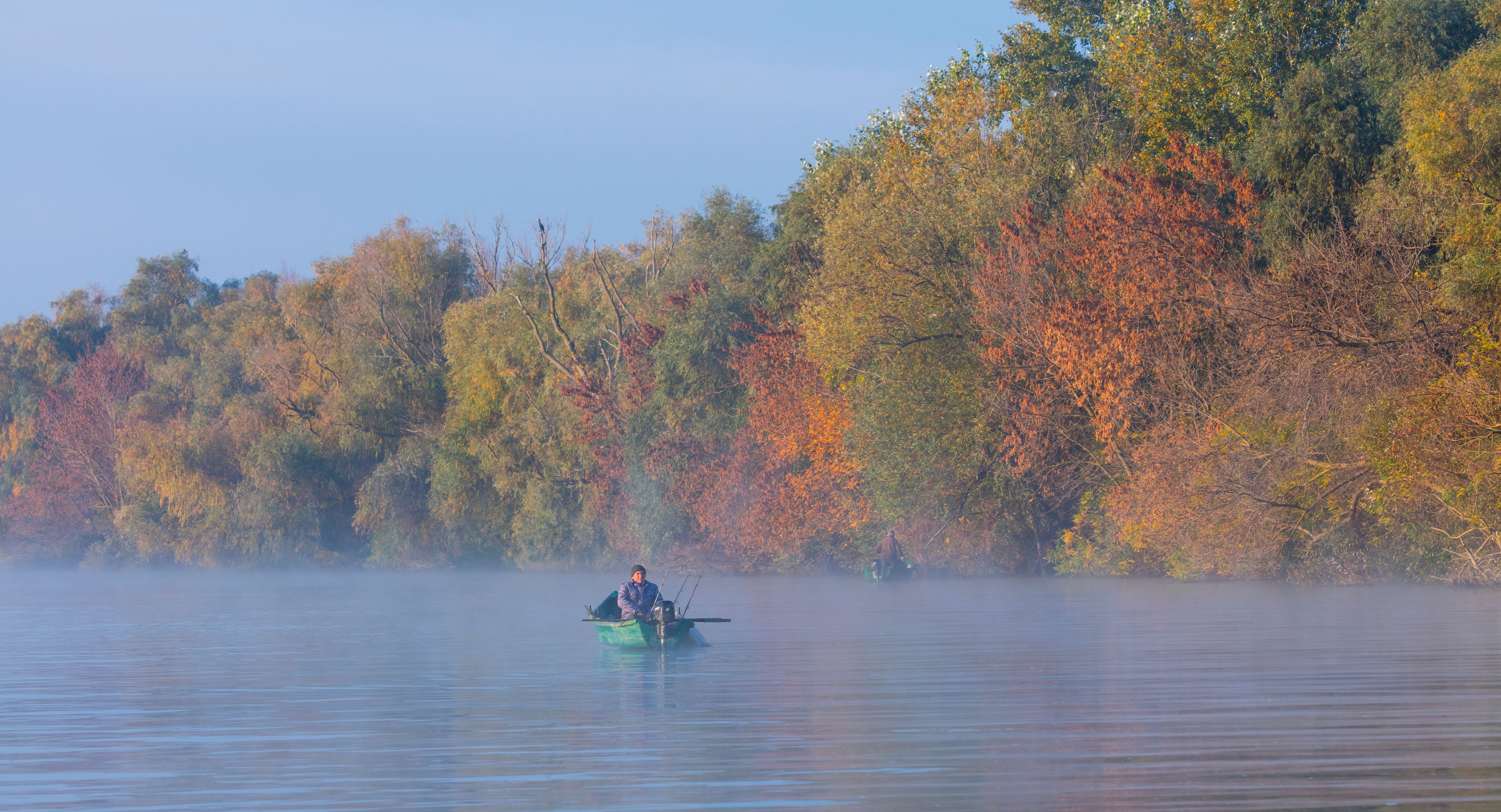 A calm lake with a person fishing in a small boat, surrounded by autumn-colored trees.