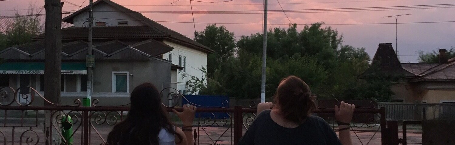 Dos personas de pie junto a un portón de metal, con vistas a una zona residencial con casas y un cielo al atardecer.