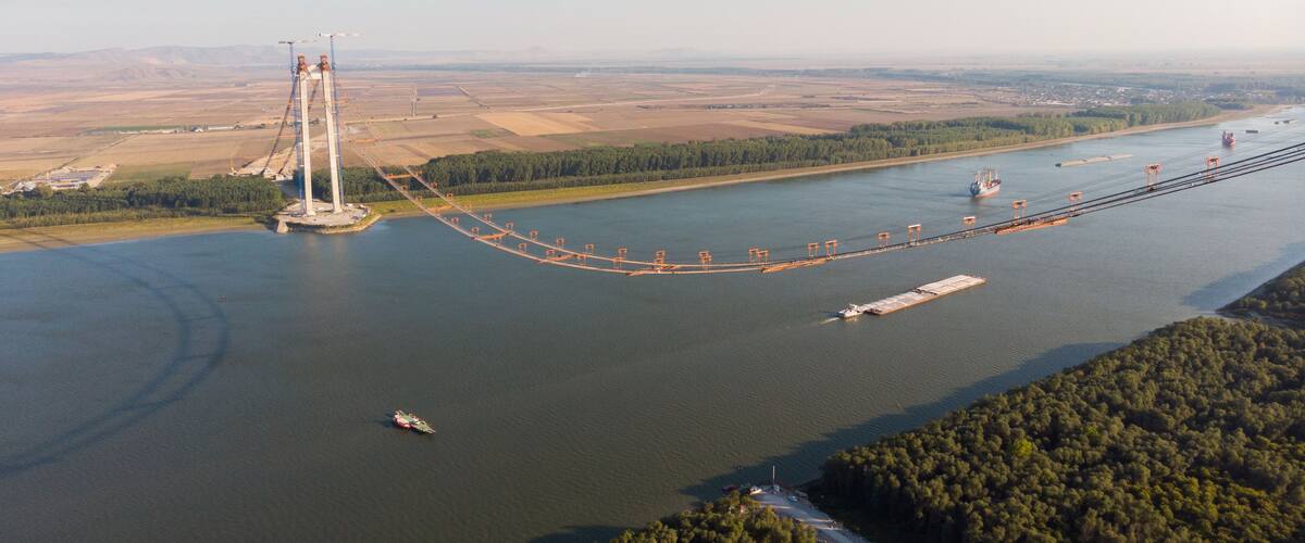 A large suspension bridge spanning a wide river, surrounded by open fields and a forested area.