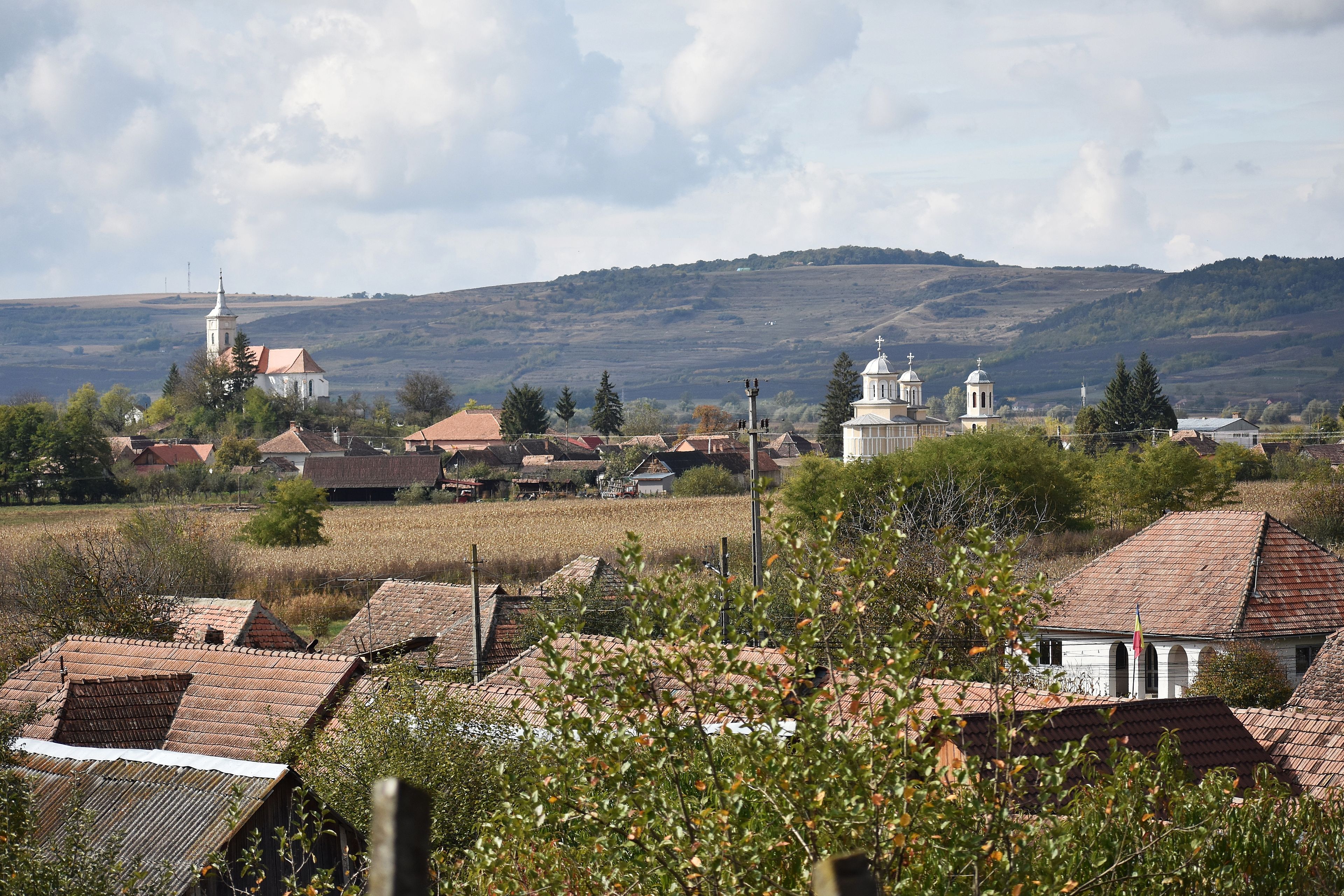 A rural landscape with a church, residential buildings, and rolling hills in the background.