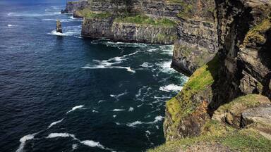 A coastal cliff with a lighthouse, ocean waves, and a grassy hill.