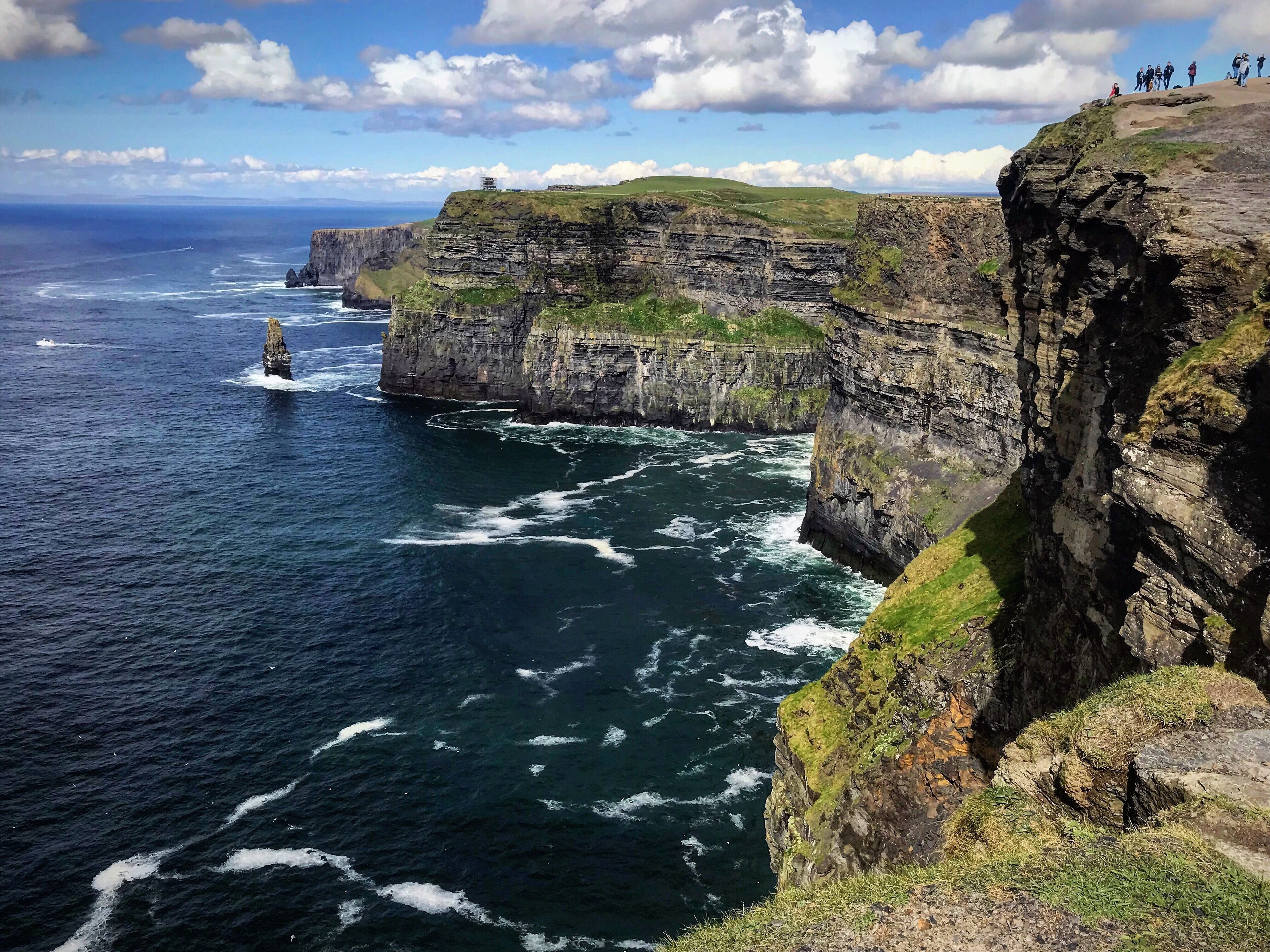 A coastal cliff with a lighthouse, ocean waves, and a grassy hill.