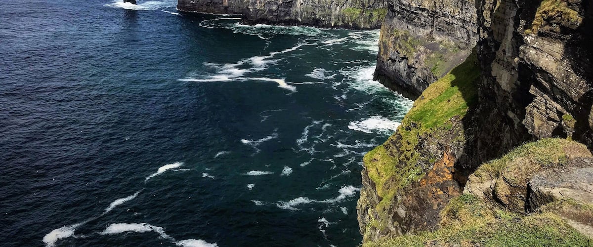 A coastal cliff with a lighthouse, ocean waves, and a grassy hill.