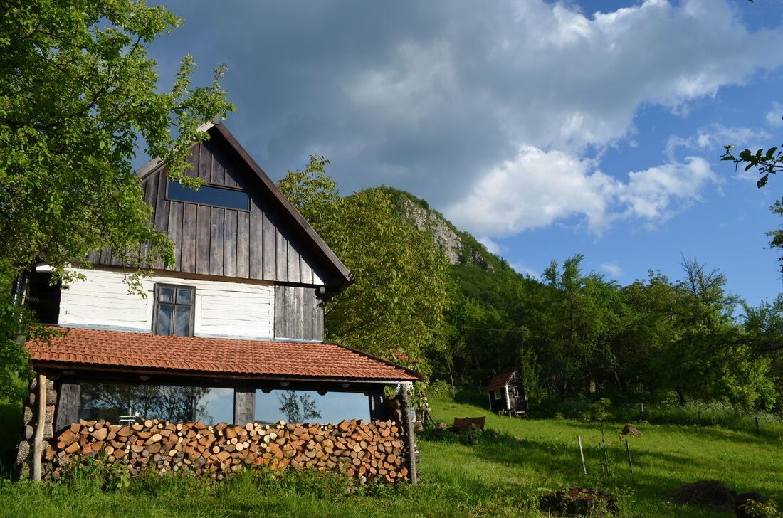 A rustic wooden house with a red-tiled roof, surrounded by lush greenery and a clear blue sky.
