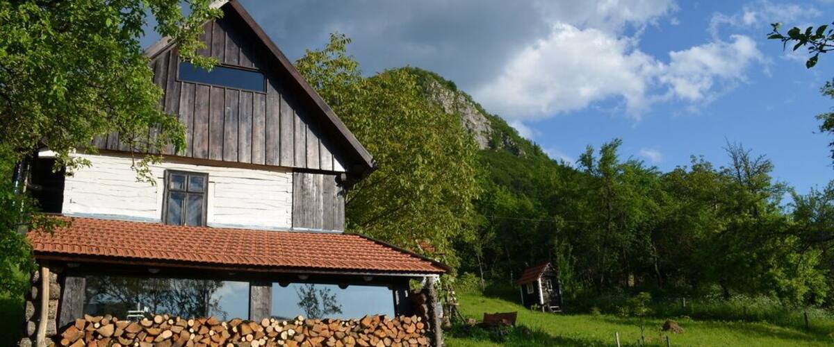 A rustic wooden house with a red-tiled roof, surrounded by lush greenery and a clear blue sky.