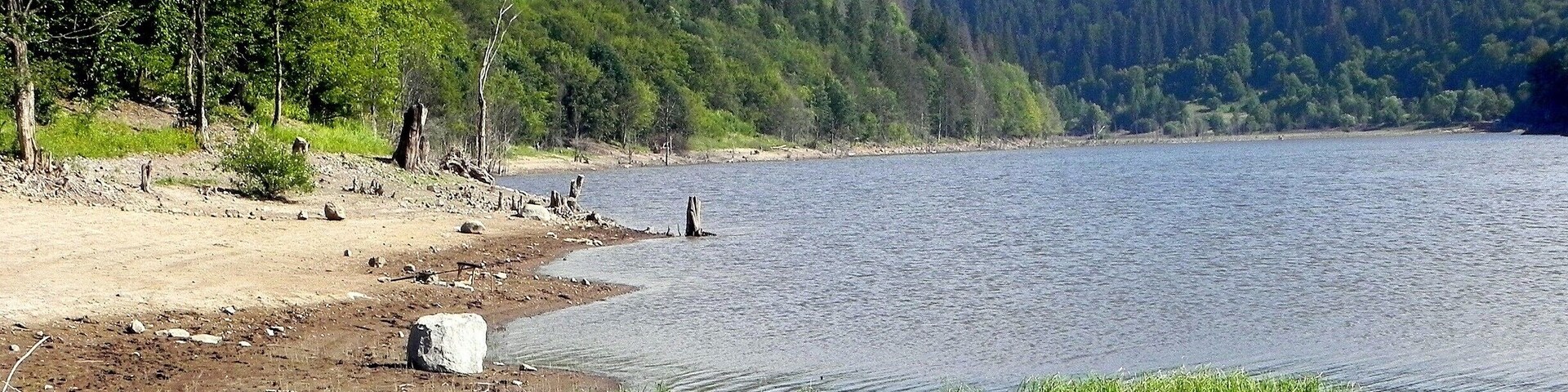 Paisagem à beira de um lago, com uma praia de areia, pedras espalhadas e colinas arborizadas ao fundo.