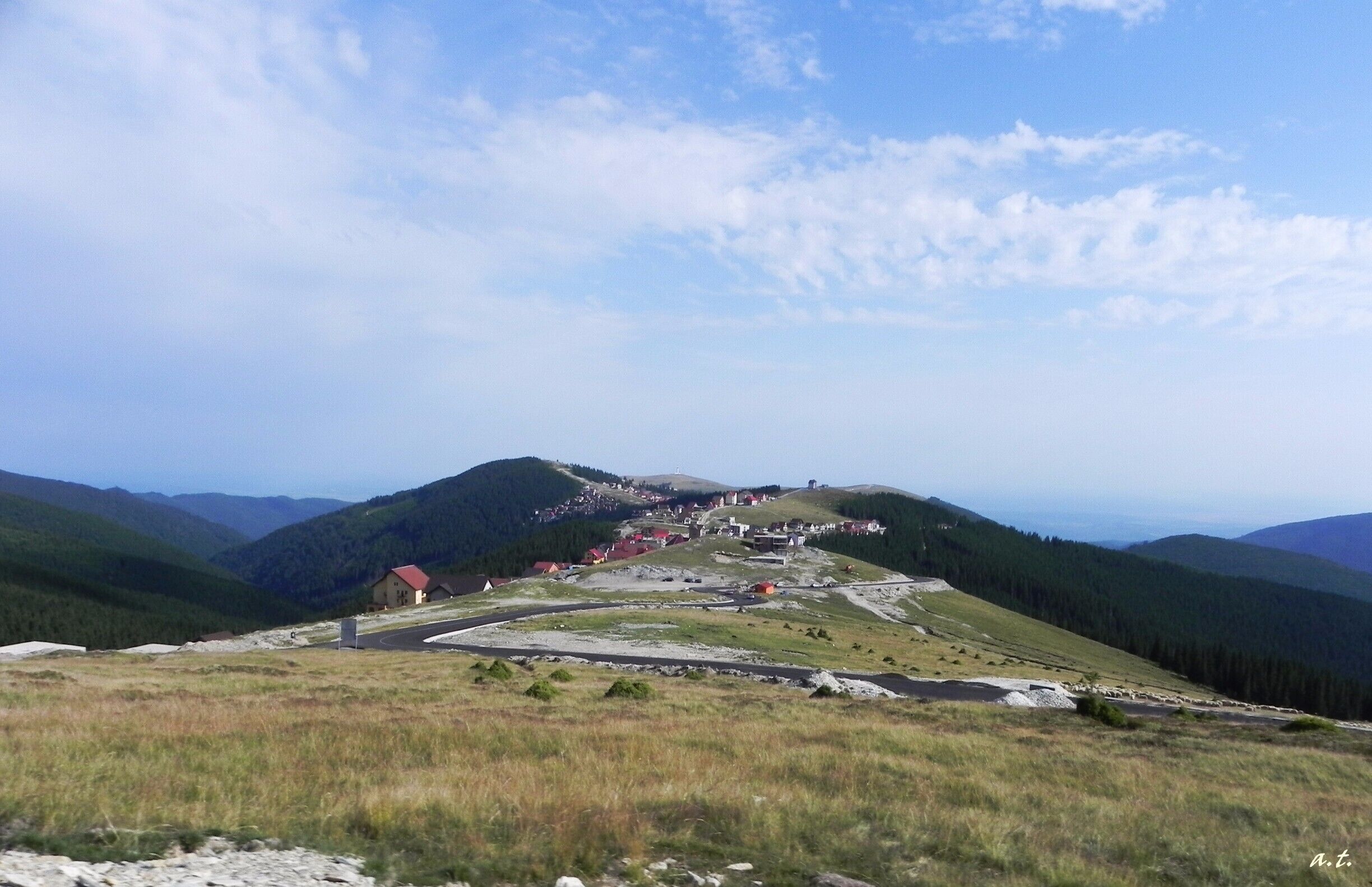 A mountainous landscape with a winding road, a small settlement, and forested hills.