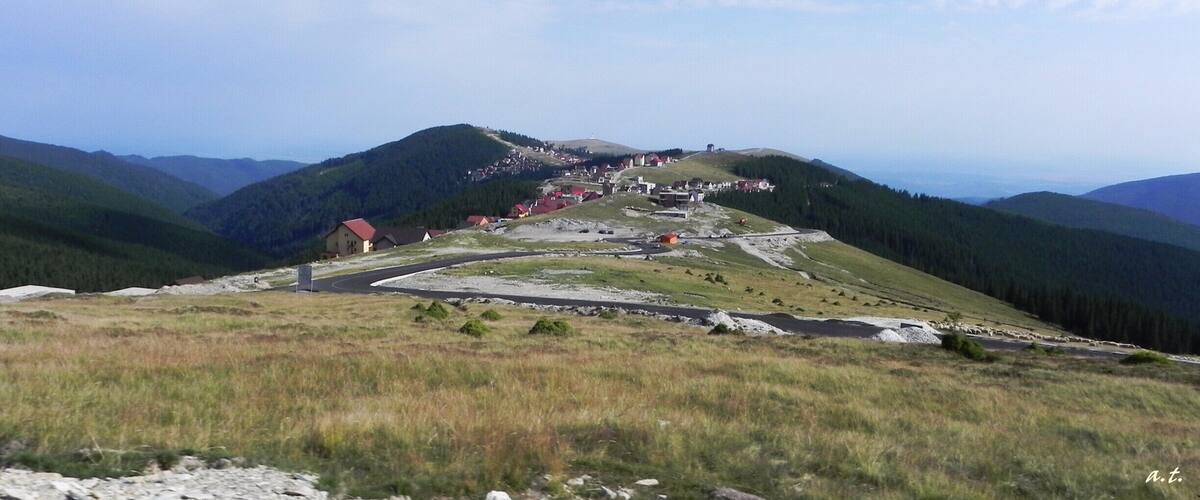 A mountainous landscape with a winding road, a small settlement, and forested hills.