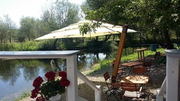 A riverside picnic area with tables and chairs, a hanging flower basket, and a large umbrella.
