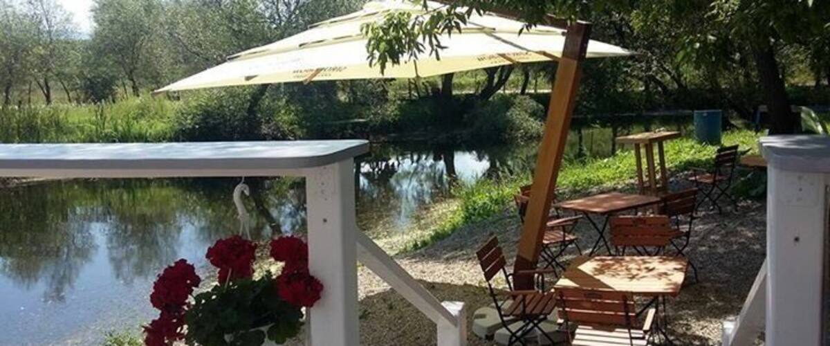 A riverside picnic area with tables and chairs, a hanging flower basket, and a large umbrella.