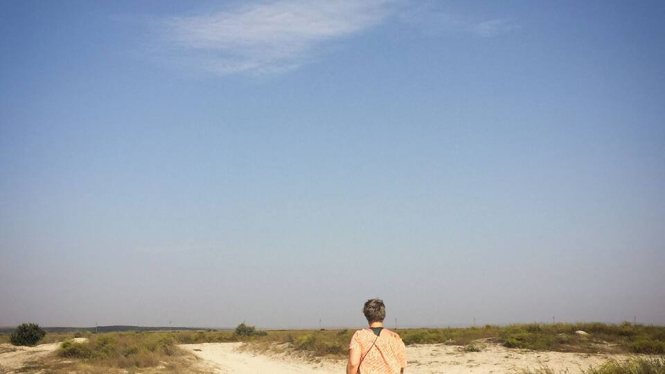 A person walking on a sandy path in a desert landscape with sparse vegetation and a clear sky.
