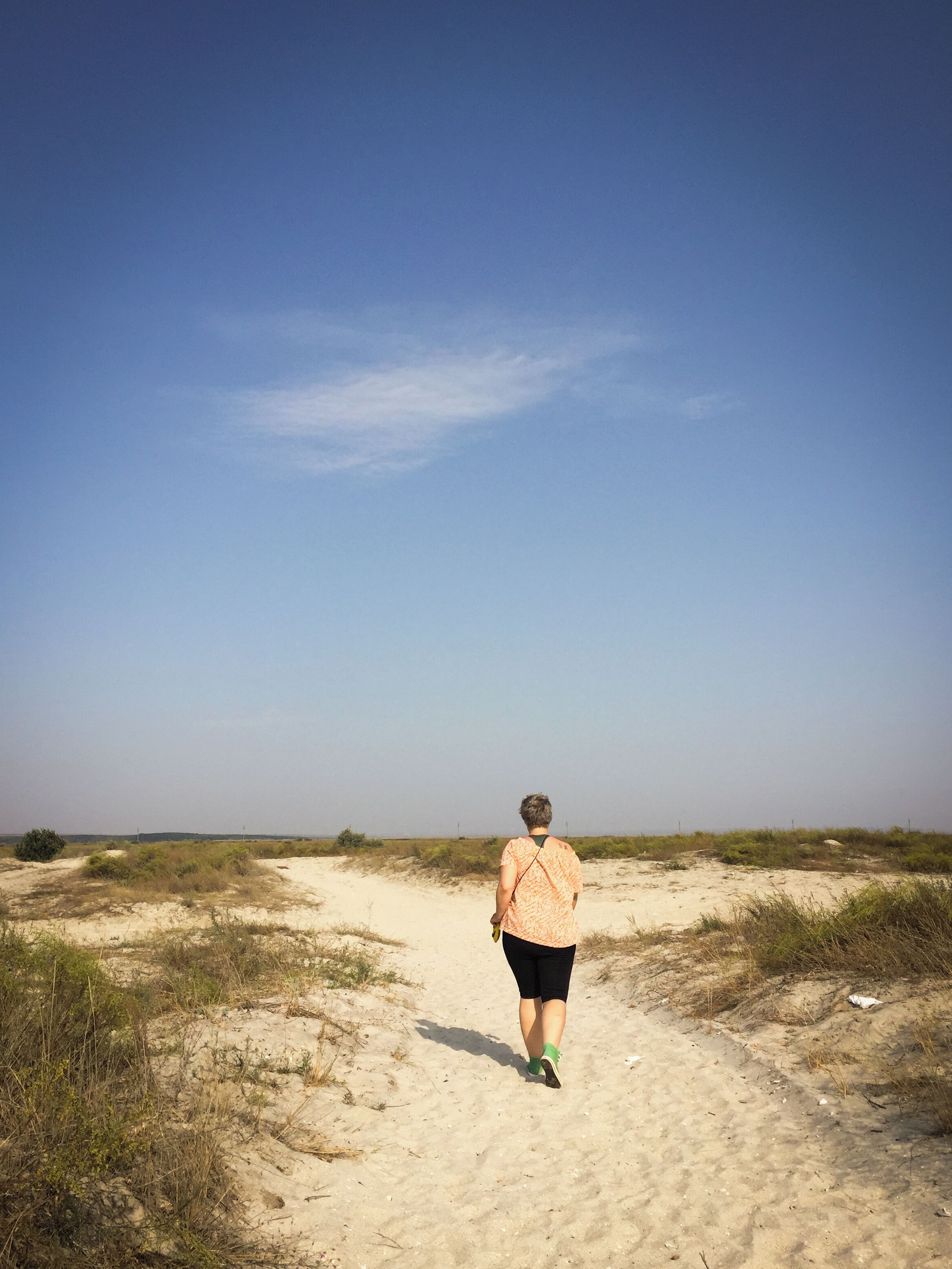A person walking on a sandy path in a desert landscape with sparse vegetation and a clear sky.