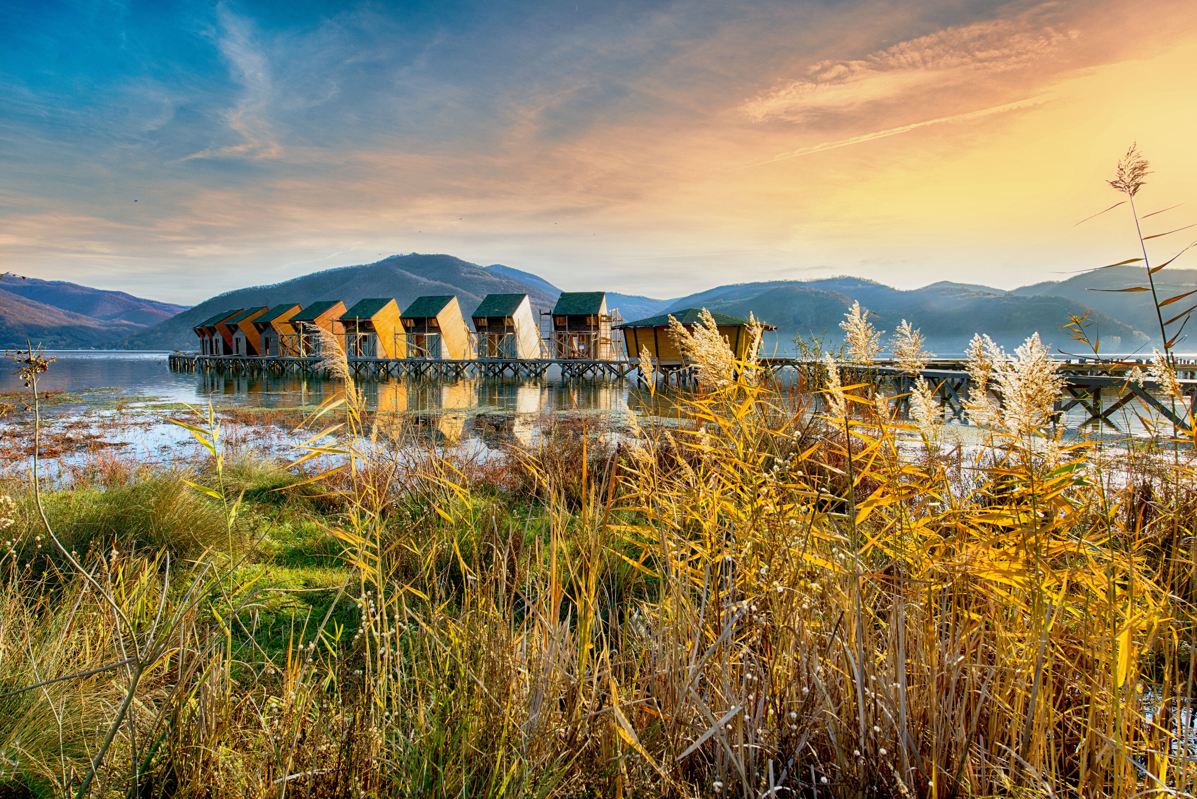 A row of wooden chalets on stilts over a lake, surrounded by mountains and tall grasses.