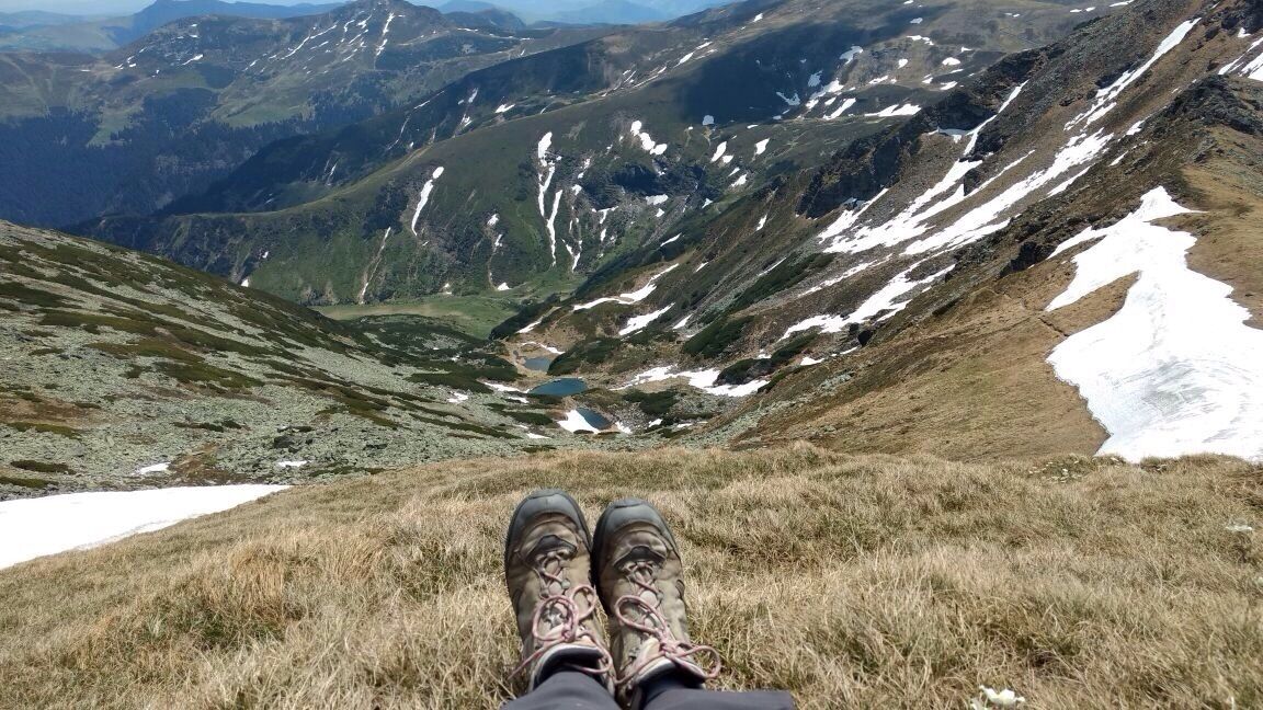 A person sitting on a grassy mountain slope with snow patches and a valley in the background.