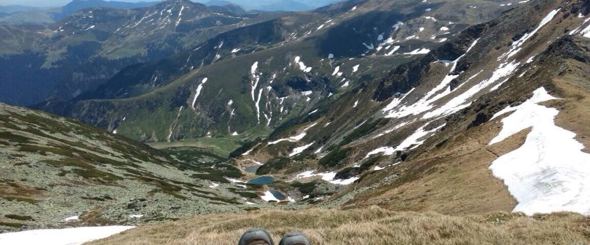 A person sitting on a grassy mountain slope with snow patches and a valley in the background.