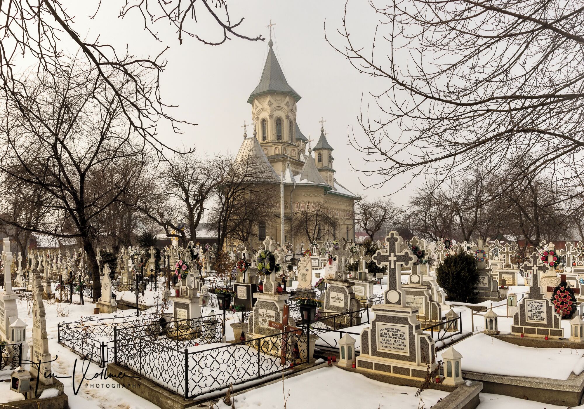 A cemetery with numerous tombstones and crosses, surrounded by leafless trees and a church with a tall spire.