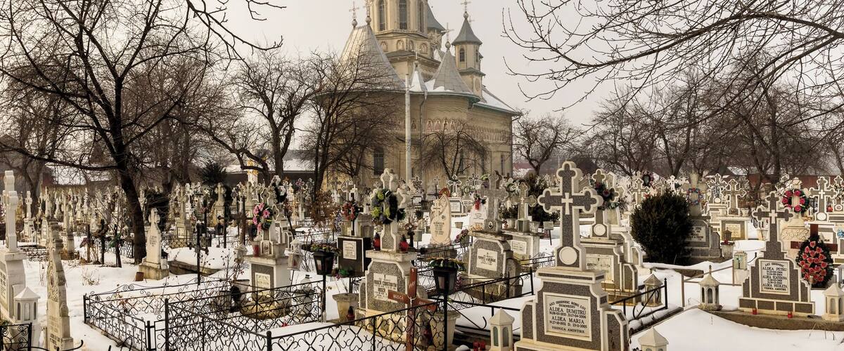 A cemetery with numerous tombstones and crosses, surrounded by leafless trees and a church with a tall spire.