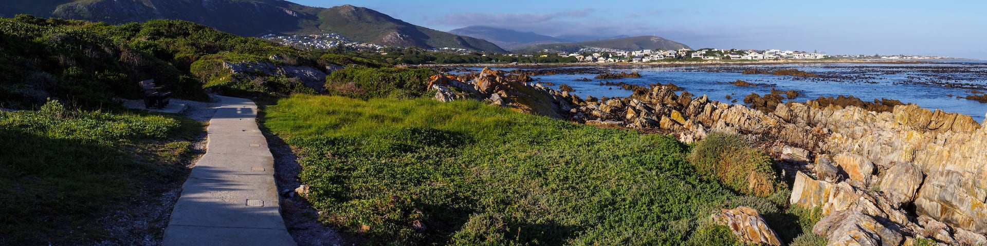 A coastal path with rocky outcrops, greenery, and a distant town.