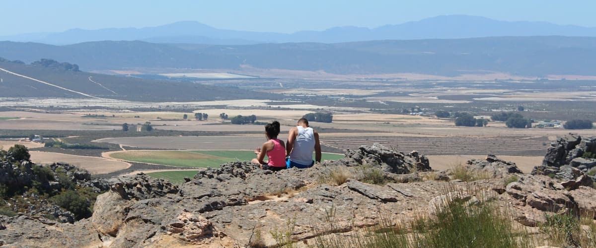 Two people sitting on a rocky outcrop overlooking a valley with fields and distant mountains.