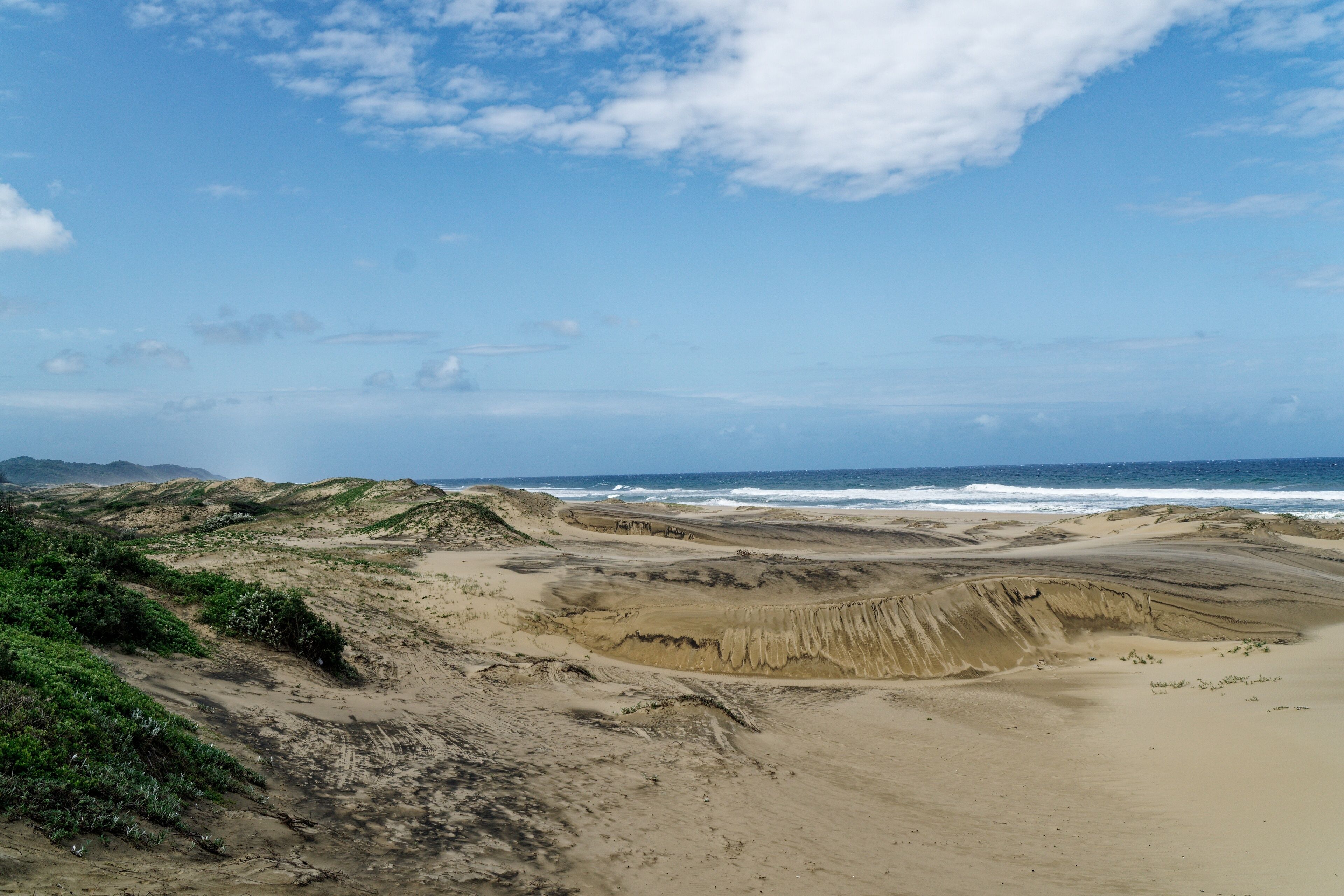 A coastal landscape with sandy terrain, green vegetation, and a distant view of the ocean.