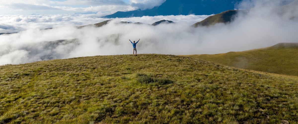 A person standing on a grassy hill with arms raised, surrounded by clouds and mountains in the background.
